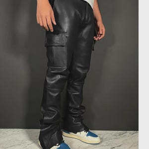 Puede incluir: Pantalones cargo de cuero negro con diseño de pierna recta. Los pantalones tienen un bolsillo cargo en la pierna izquierda y se combinan con zapatillas deportivas azules y blancas. Los pantalones son de un material liso y brillante.
