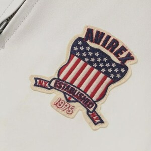 K&ouml;nnte beinhalten: Wei&szlig;e Lederjacke mit aufgen&auml;htem Aufn&auml;her. Der Aufn&auml;her ist schildf&ouml;rmig und zeigt das Design der amerikanischen Flagge mit roten und wei&szlig;en Streifen und wei&szlig;en Sternen auf blauem Grund. Der Aufn&auml;her lautet "AVIREX N.Y. ESTABLISHED 1975".