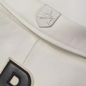 K&ouml;nnte beinhalten: Nahaufnahme eines wei&szlig;en Lederartikels mit einer Naht. Ein kleiner, gestickter Aufn&auml;her mit einem stilisierten Design ist sichtbar. Ein gr&ouml;&szlig;erer Aufn&auml;her mit schwarzem Rand und dem Buchstaben "D" ist ebenfalls vorhanden.