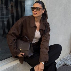 May include: A woman wearing brown sunglasses, a brown suede jacket, a white top, black pants, and black loafers. She is holding a brown handbag with a top handle. A gold watch is on her wrist.