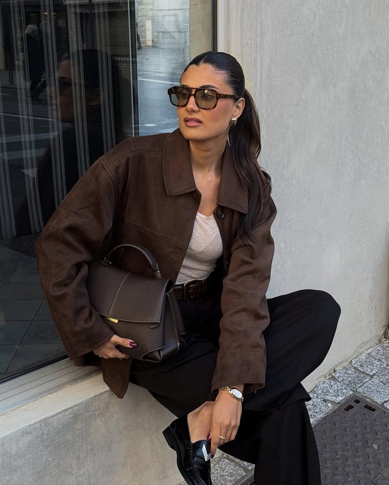 May include: A woman wearing brown sunglasses, a brown suede jacket, a white top, black pants, and black loafers. She is holding a brown handbag with a top handle. A gold watch is on her wrist.