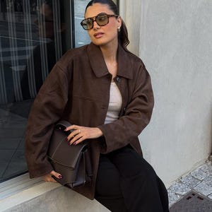 May include: A woman wearing brown sunglasses, a brown suede jacket, a light gray top, and black pants. She is holding a dark brown leather handbag. The background includes a window and a concrete wall.