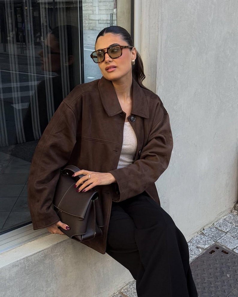 May include: A woman wearing brown sunglasses, a brown suede jacket, a light gray top, and black pants. She is holding a dark brown leather handbag. The background includes a window and a concrete wall.