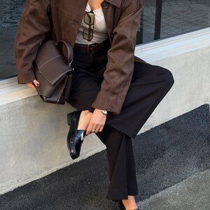 May include: A brown suede jacket, a dark brown handbag, and black loafers with a silver accent. The outfit includes a gray top, black pants, and a brown belt. Sunglasses are tucked into the top.