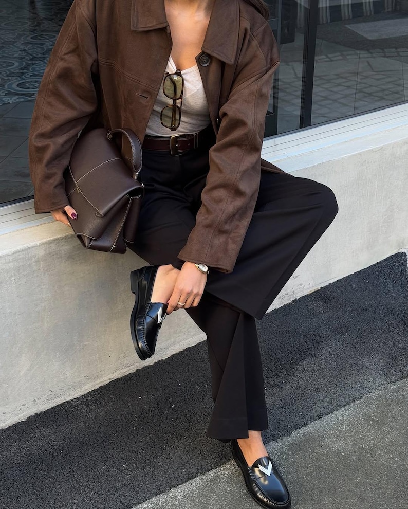 May include: A brown suede jacket, a dark brown handbag, and black loafers with a silver accent. The outfit includes a gray top, black pants, and a brown belt. Sunglasses are tucked into the top.