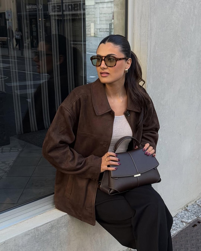 May include: A brown suede jacket with a button-up front. The image also shows a brown leather handbag with a silver clasp, black pants, and brown-framed sunglasses.