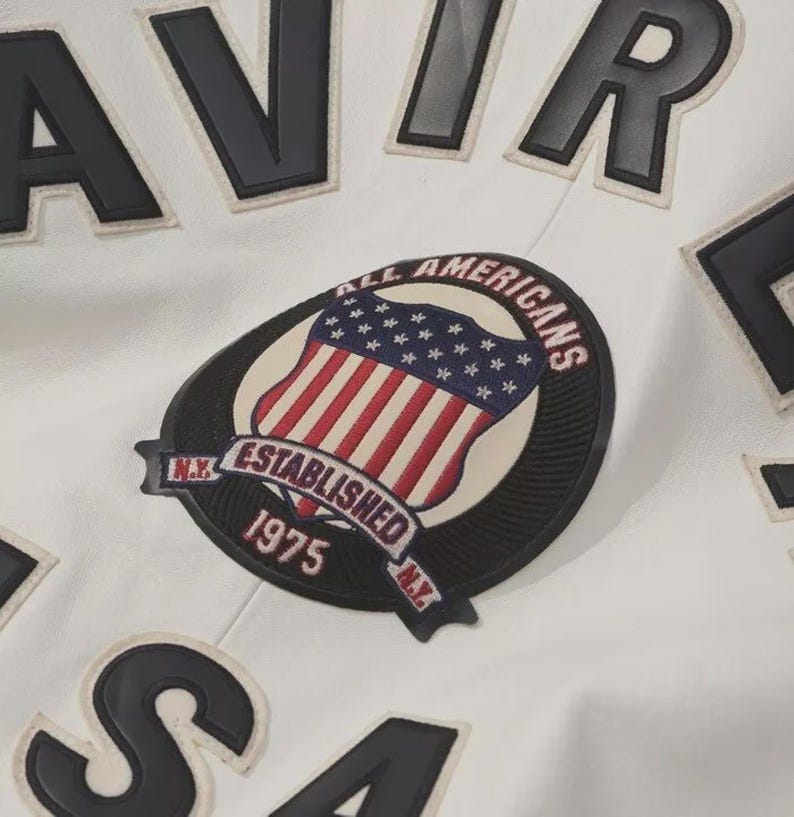 K&ouml;nnte beinhalten: Nahaufnahme einer wei&szlig;en Jacke mit schwarzen Buchstaben und Aufn&auml;hern. Die Jacke hat einen Aufn&auml;her mit einem amerikanischen Flaggen-Design und dem Text "ALL AMERICANS ESTABLISHED 1975 N.Y."