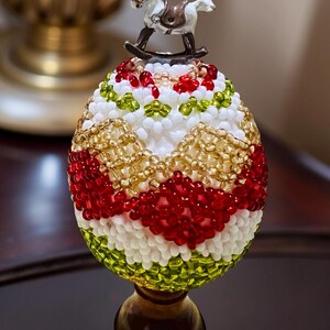 May include: An egg-shaped ornament decorated with white, red, gold, and green beads. A small rocking horse figurine is placed on top. The ornament is mounted on a dark wooden base, creating a detailed and festive design.