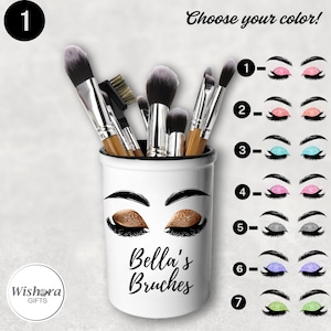 Può includere: Portapennelli da trucco in ceramica bianca con bordo nero. Il portapennelli ha un design con un paio di occhi con ombretto glitter oro e ciglia nere. Il testo "Bella's Brushes" è stampato sotto gli occhi. Il portapennelli è pieno di vari pennelli da trucco.