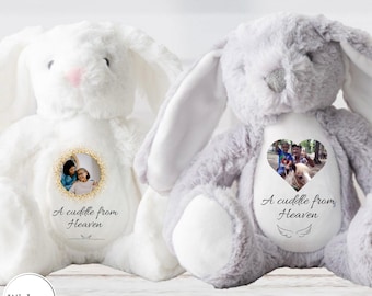 May include: Two plush stuffed rabbits, one white and one grey, each featuring a personalised circular or heart-shaped photo patch on the chest. The patches include the text A cuddle from Heaven and small line drawings of angel wings.