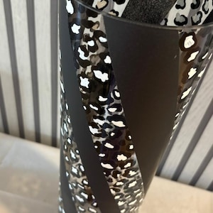 Puede incluir: Un jarrón alto y cilíndrico con exterior negro y un patrón decorativo. El patrón incluye rayas negras y secciones con un diseño de estampado de leopardo en blanco, negro y beige. El jarrón es de vidrio.
