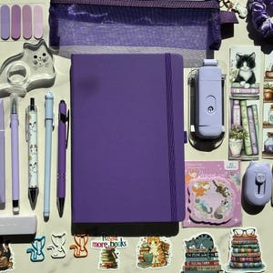 May include: A purple notebook centered among stationery items including a mesh pencil case, cat-shaped tape dispenser, various pens, sticky notes, bookmarks, and stickers featuring cats and books. The collection includes a purple scrunchie and a small portable light.
