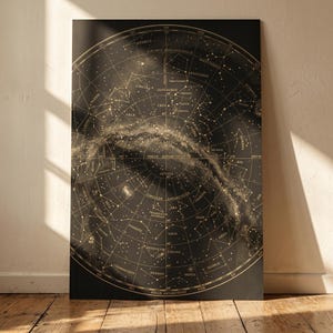 Op de afbeelding: Een vintage-stijl, cirkelvormige sterrenkaart print. De print heeft een donkere achtergrond met een gedetailleerde kaart van sterrenbeelden en hemellichamen in goud. De print wordt weergegeven op een houten vloer.