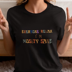 Op de afbeelding: Zwart T-shirt met de tekst "EVERYONE RELAX I'M MOSTLY SANE" in kleurrijke, retro-stijl letters. De persoon draagt het shirt en maakt een vredesteken met hun vingers.