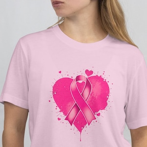 Op de afbeelding: Lichtroze T-shirt met een roze lintontwerp in een hartvorm. Het ontwerp bevat een roze lint en kleine hartjes. Het shirt is gemaakt van een zacht materiaal en heeft korte mouwen.