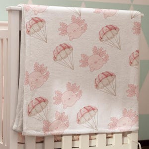Op de afbeelding: Een wit babybedje met een roze en wit babydekentje met een patroon van roze axolotls met parachutes. Het bedje staat in een kamer met een lichtgroen en wit geometrisch behang.