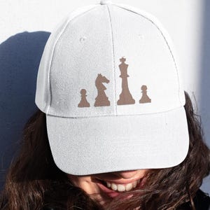 Puede incluir: Gorra de béisbol blanca con un diseño de piezas de ajedrez marrones. El diseño incluye un peón, un caballo, un rey y otro peón. La gorra está hecha de tela clara y tiene una visera curva.