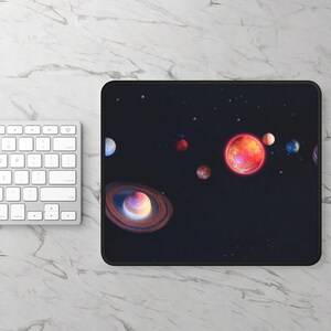 Op de afbeelding: Een rechthoekige zwarte muismat met een levendige illustratie van planeten in een sterrenhemel. De planeten zijn afgebeeld in verschillende kleuren, waaronder rood, oranje, blauw en bruin, met de ringen van Saturnus prominent aanwezig. Een wit toetsenbord is zichtbaar op de achtergrond.
