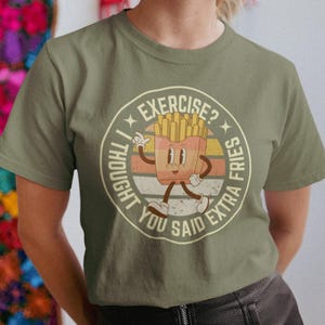 Puede incluir: Una camiseta verde con un gráfico retro de un personaje de papas fritas de dibujos animados. El gráfico dice "Pensé que habías dicho papas fritas extra" y "¿ejercicio?" en un círculo.