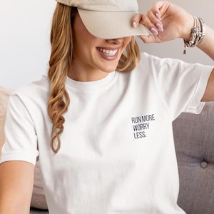 Könnte beinhalten: Weißes T-Shirt mit dem Text "RUN MORE WORRY LESS" in Marineblau. Eine beige Baseballkappe wird getragen. Die Person lächelt. Das Bild zeigt eine Person, die ein T-Shirt und eine Kappe trägt.