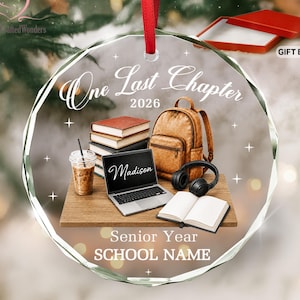 May include: A round glass ornament featuring a red ribbon, printed with the text One Last Chapter 2026, Senior Year, and SCHOOL NAME. The design shows a laptop labeled Madison, a stack of books, a backpack, headphones, and a coffee cup.