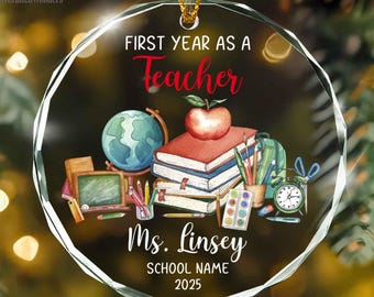 May include: A circular glass ornament featuring the text FIRST YEAR AS A Teacher, Ms. Linsey, SCHOOL NAME, and 2025. The design includes a globe, a stack of books topped with a red apple, a pencil holder, a paint set, and a clock with scissors.