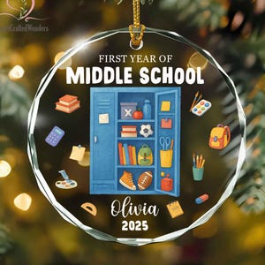 May include: A circular clear glass ornament featuring the text FIRST YEAR OF MIDDLE SCHOOL, Olivia, and 2025. The design includes a blue school locker filled with books, a soccer ball, and a backpack, surrounded by school supplies like a calculator.