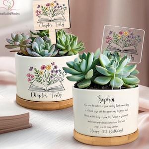 May include: Two white ceramic succulent planters with bamboo bases. Each planter features a floral and butterfly design with the text "Chapter Forty." One planter has a wooden sign, the other a clear sign. One planter has the text "Sophia" and a birthday message.