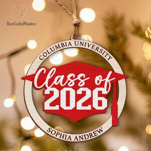 May include: Wooden graduation ornament with the text "Columbia University" and "Class of 2026". The ornament has a red mortarboard design and the name "Sophia Andrew" at the bottom. It is hanging from a natural twine string.