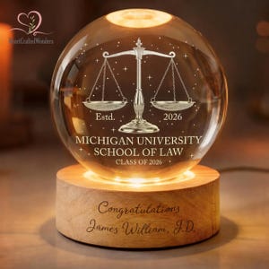 May include: A lit glass sphere on a wooden base, engraved with a scale of justice, the text "Estd. 2026", and "MICHIGAN UNIVERSITY SCHOOL OF LAW CLASS OF 2026". The base reads "Congratulations James William, J.D."
