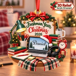 May include: A 3D relief acrylic Christmas ornament featuring a desk scene with a laptop displaying the name Daniel, a red Santa hat, a green desk lamp, books, a red alarm clock, headphones, and a scarf. The sign reads Freshman Year Christmas 2026.