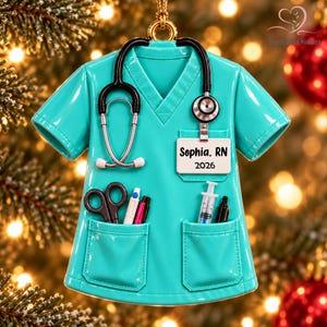 May include: A teal scrub top ornament featuring a black stethoscope draped around the neck, a white ID badge printed with "Sophia, RN 2026", and two front pockets containing black scissors, a white pen, a pink pen, a syringe, and a black pen.