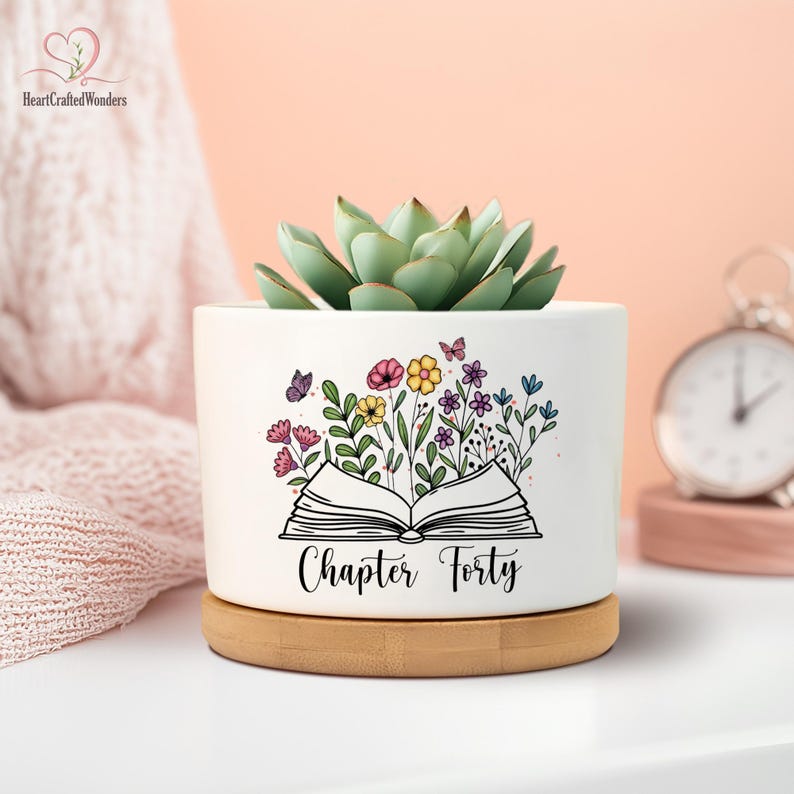 May include: A white ceramic planter with a succulent plant. The planter features a floral design with butterflies and the text "Chapter Forty." It sits on a wooden saucer.