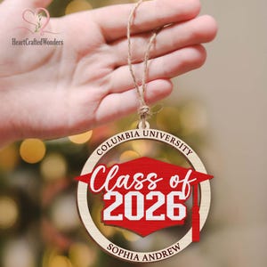 May include: Wooden Columbia University graduation ornament. The ornament features a red graduation cap and the text "Class of 2026" in white. The name "Sophia Andrew" is also included. The ornament is suspended by a twine string.