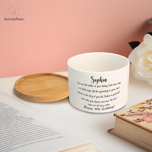 May include: A white ceramic planter with a wooden coaster. The planter has a personalized message for Sophia, wishing her a happy 40th birthday. The text is in black script. A book and a white flower are in the background.