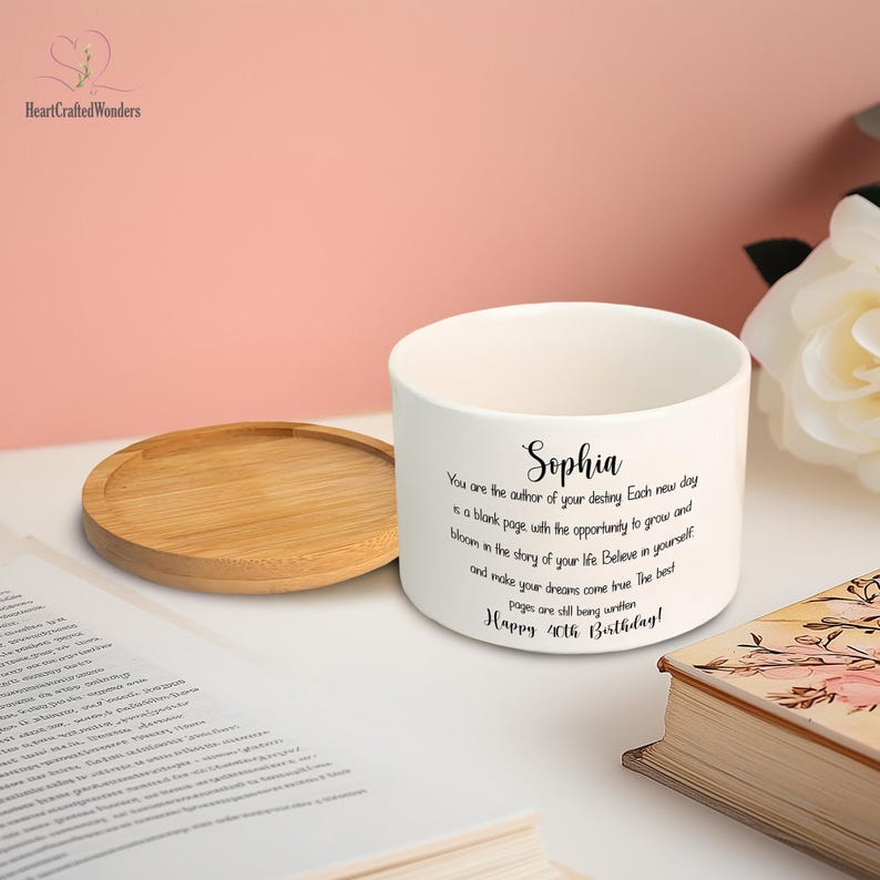 May include: A white ceramic planter with a wooden coaster. The planter has a personalized message for Sophia, wishing her a happy 40th birthday. The text is in black script. A book and a white flower are in the background.