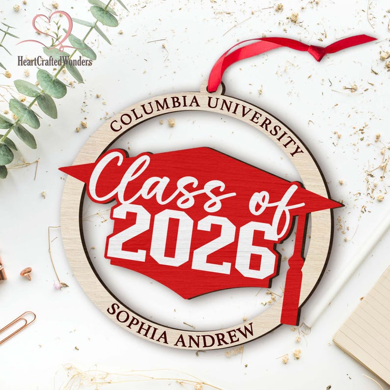 May include: A wooden graduation ornament with a red mortarboard design. The ornament is circular, with the words "Columbia University," "Class of 2026," and "Sophia Andrew" inscribed. A red ribbon is attached for hanging.