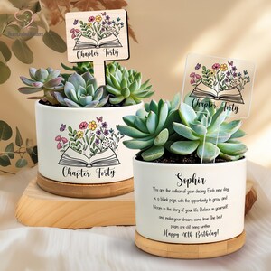 May include: Two white ceramic succulent planters with wooden bases. Each planter features a floral design with an open book and the text "Chapter Forty." One planter has a clear sign that says "Chapter Forty."