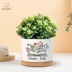 May include: A white ceramic planter with a green plant, featuring a floral and butterfly design with the text "Chapter Forty." The planter sits on a round wooden base, creating a decorative accent.