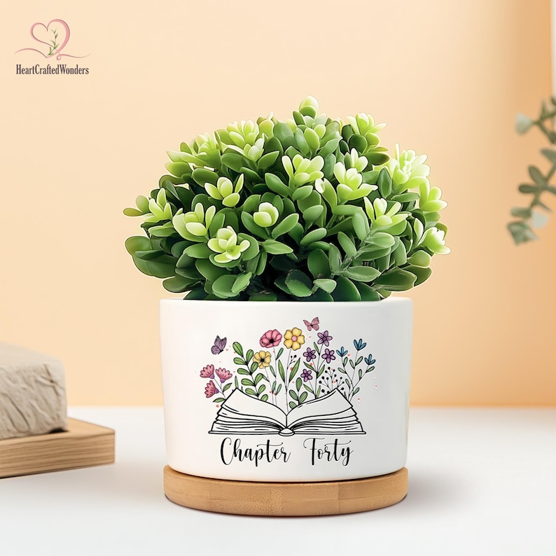 May include: A white ceramic planter with a green plant, featuring a floral and butterfly design with the text "Chapter Forty." The planter sits on a round wooden base, creating a decorative accent.