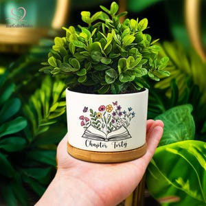 May include: A small, white ceramic planter with a floral and butterfly design, and the text "Chapter Forty." The planter holds a vibrant green plant and sits on a bamboo base. The background is a lush display of green foliage.
