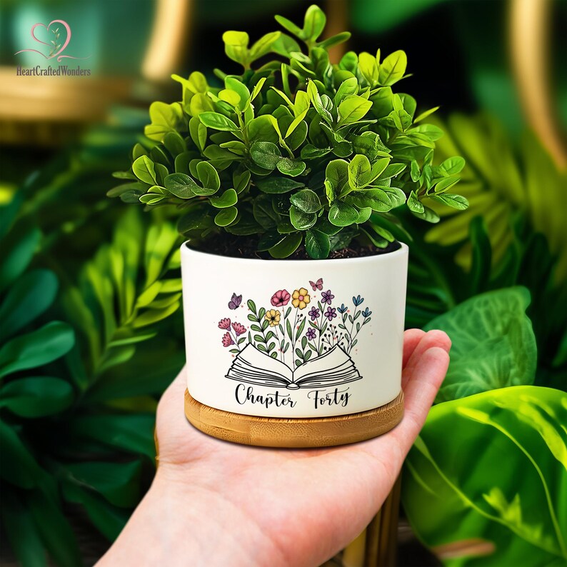 May include: A small, white ceramic planter with a floral and butterfly design, and the text "Chapter Forty." The planter holds a vibrant green plant and sits on a bamboo base. The background is a lush display of green foliage.