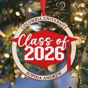 May include: A wooden Christmas ornament with a red graduation cap design. The ornament features the text "Columbia University", "Class of 2026", and "Sophia Andrew". It is hanging from a red ribbon against a blurred background of Christmas lights.