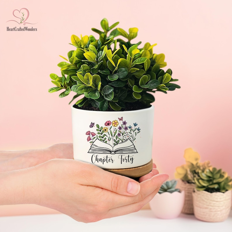 May include: A white ceramic planter with a floral design and the text "Chapter Forty" holds a green plant. The planter sits on a wooden base. The background is a soft pink.