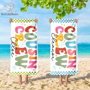 May include: Two white beach towels with the words "Cousin Crew" in colourful retro font. One towel says "Chloe" and the other says "Eleanor". Each towel has a checkered border and is held up on a beach.