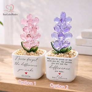 May include: Two decorative glass flower arrangements in white square pots. One is pink, the other purple. Each pot has a message and the name "Olivia". The arrangements sit on a bed of small stones.