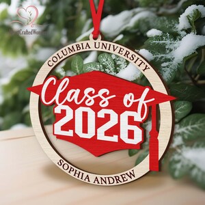 May include: A wooden Columbia University ornament with a red graduation cap design. The ornament reads "Class of 2026" and "Sophia Andrew" in white script. The ornament hangs from a red ribbon.