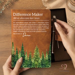 May include: A brown leather journal with the words "Difference Maker" and a definition printed on the cover. The journal features a watercolor forest design at the bottom. A pen is included in a gift box.