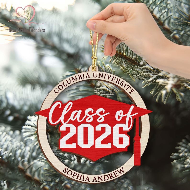 May include: A wooden graduation ornament with the text "Columbia University Class of 2026" and the name "Sophia Andrew" in a circular design. The ornament features a red graduation cap and a gold string for hanging.