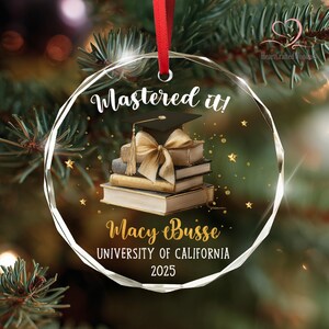 May include: Clear, round graduation ornament with a red ribbon. The ornament features the text "Mastered it!" and a stack of books with a graduation cap and bow. The name "Macy Buisse" and "UNIVERSITY OF CALIFORNIA 2025" are also displayed.